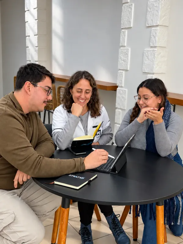 Tres personas alrededor de una mesa miran una tablet y sonríen, con un cuaderno de Stratio sobre la mesa